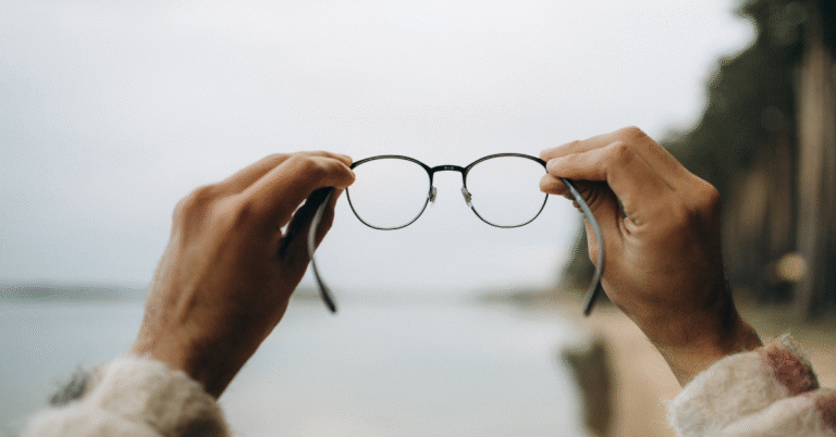 Hands holding eyeglasses toward a blurred background, symbolizing clarity, focus, and building a business anchored in vision instead of pressure.
